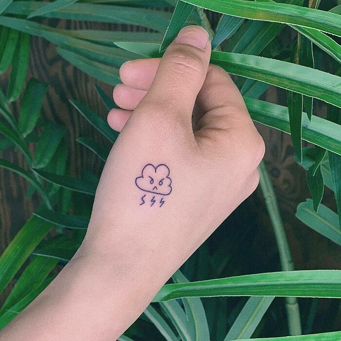 Small hand tattoo of an angry cloud with lightning bolts, surrounded by green leaves.