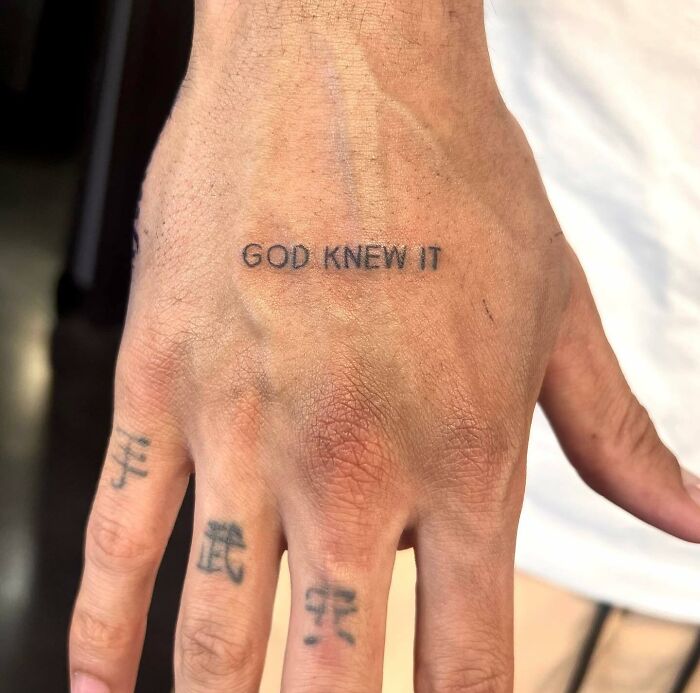 Small hand tattoo with text "God Knew It" and symbols on fingers.