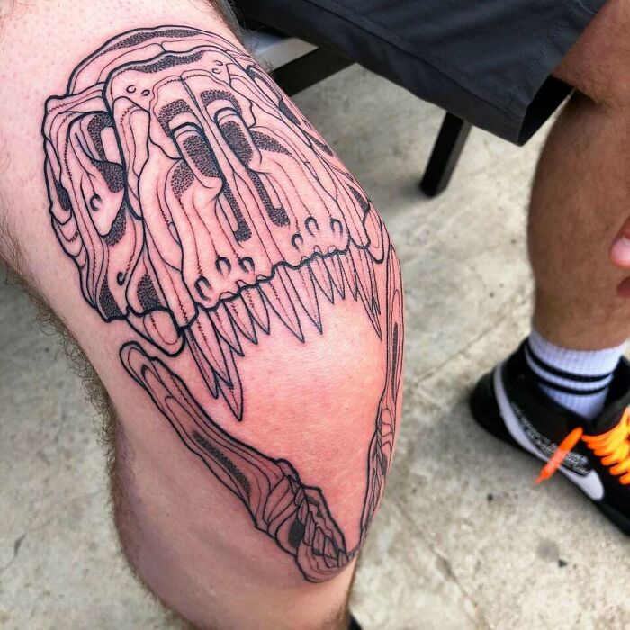 Knee tattoo of a dinosaur skull on a person's leg, showcasing bold linework in a modern design.
