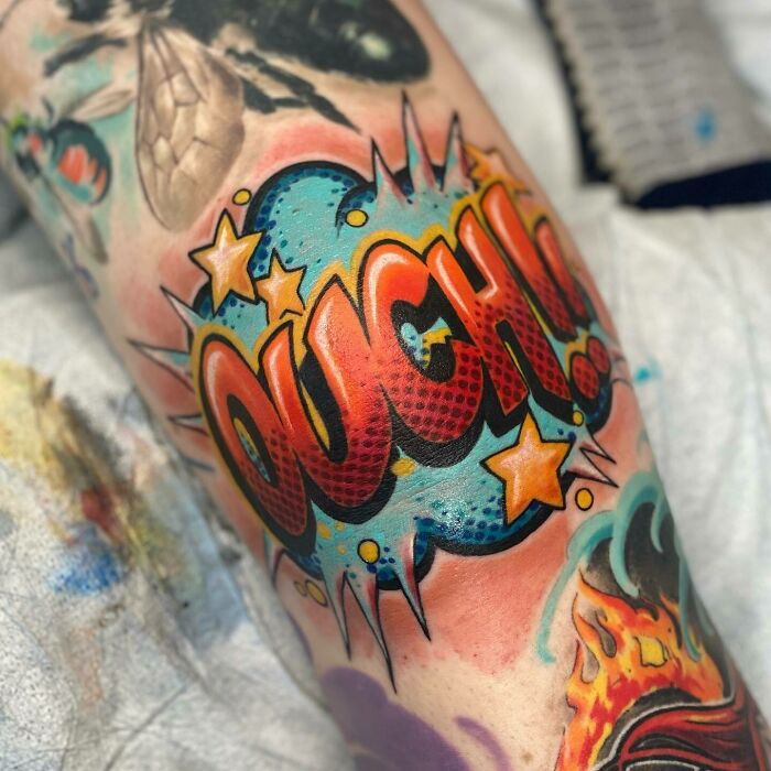 Colorful comic-style knee tattoo with the word "OUCH" in bold letters and vibrant stars.