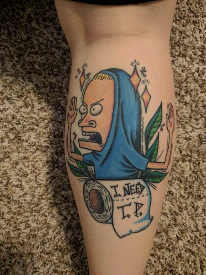 '90s tattoo featuring a cartoon character wrapped in a blue cloak holding a sign reading "I need T.P."