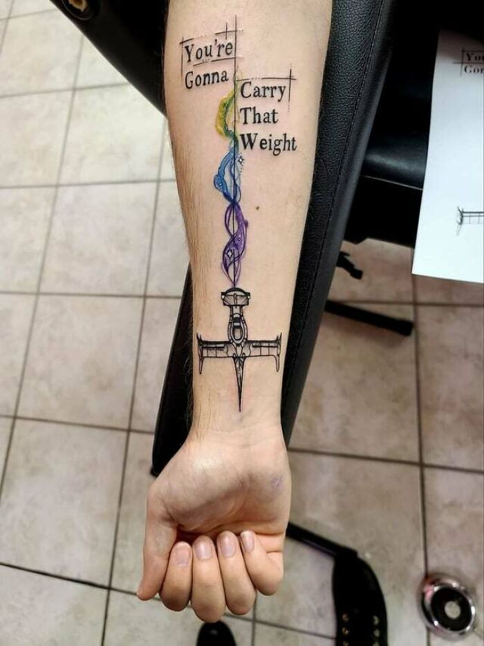 Tattoo of a sword with colorful design on arm, featuring '90s cultural phrase "You're Gonna Carry That Weight."