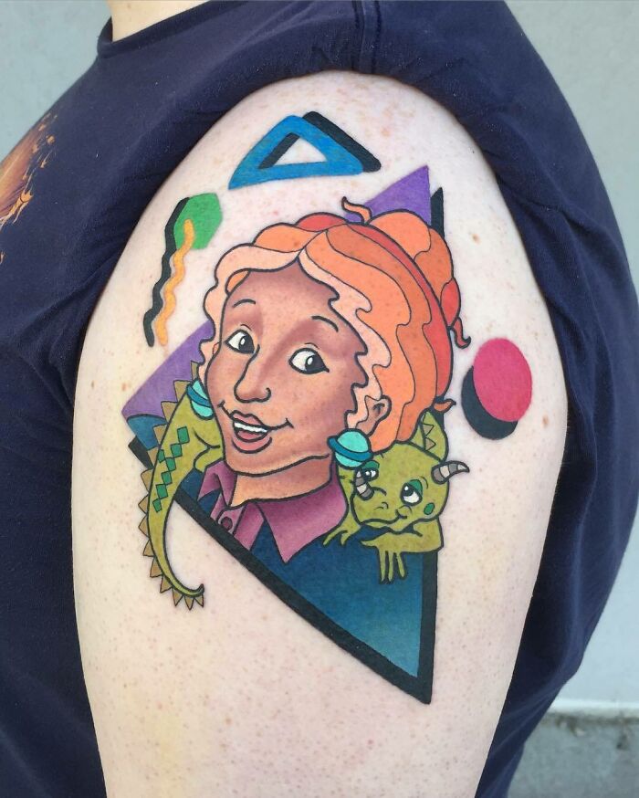 '90s tattoo featuring cartoon girl and cute creature on shoulder, colorful geometric shapes in background.