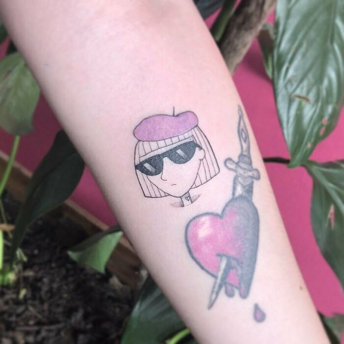 Tattoo featuring a ‘90s style cartoon character with sunglasses, a pink beret, and a heart pierced by a dagger.