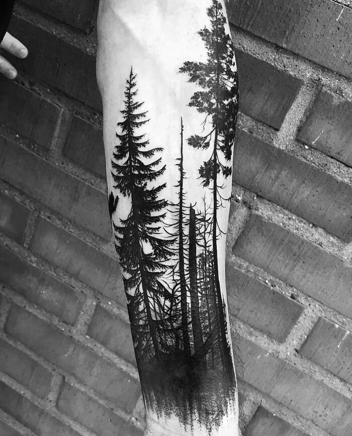 Majestic tree tattoo on an arm, depicting tall, detailed trees against a brick wall backdrop.