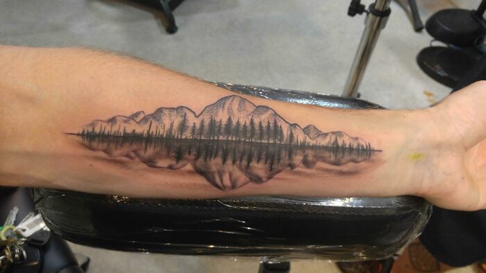 Arm with a detailed tree tattoo featuring mountains and reflections, depicting nature's wonders.
