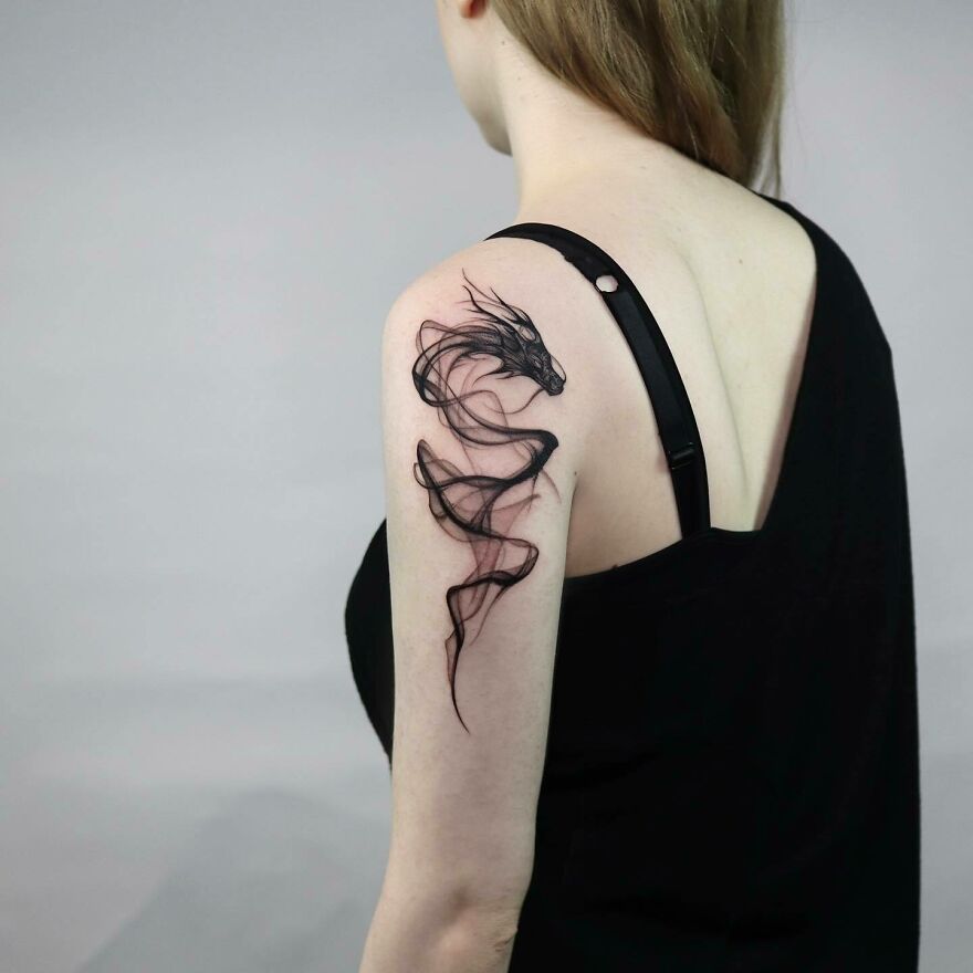 Woman with a delicate dragon tattoo on her upper arm, featuring a sleek, minimalist design in black ink.