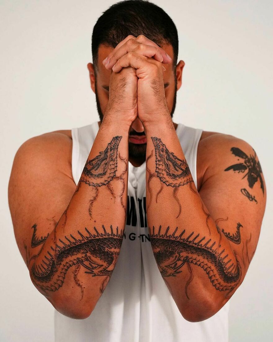 Man with dragon tattoos on his arms, hands clasped, wearing a white tank top.
