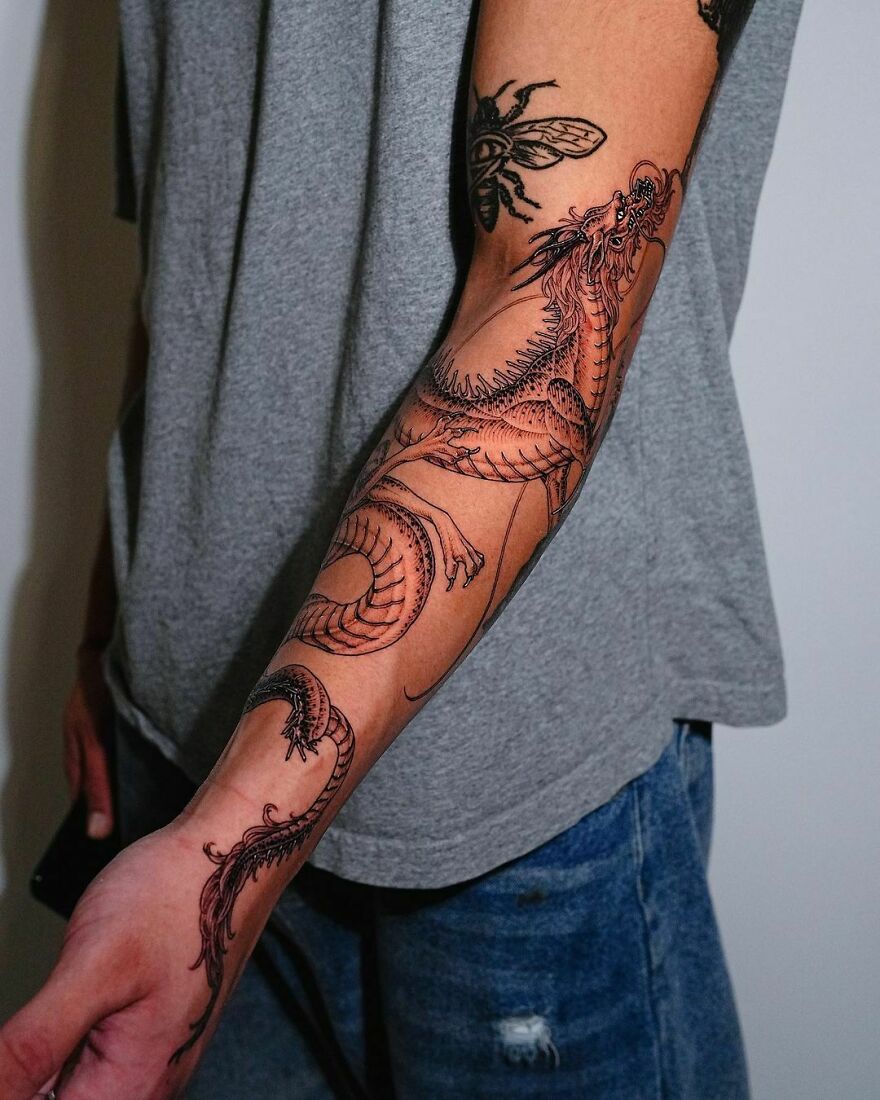 Arm with a detailed dragon tattoo in black ink, wearing a gray shirt and jeans.