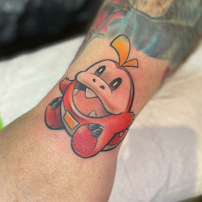 Cute anime tattoo of a cartoon dinosaur in vibrant red and orange hues on a forearm, showcasing anime tattoo art.