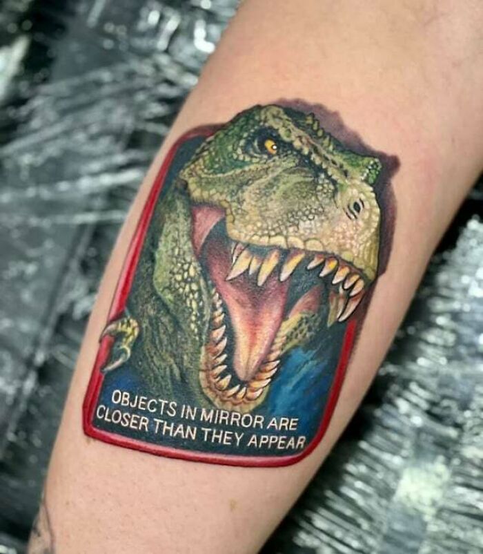 '90s tattoo of a roaring T-Rex with "Objects in mirror are closer than they appear" text on arm.