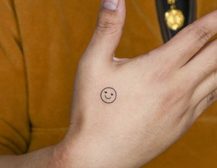 Small hand tattoo of a smiley face in black ink on the side of a thumb against a brown background.