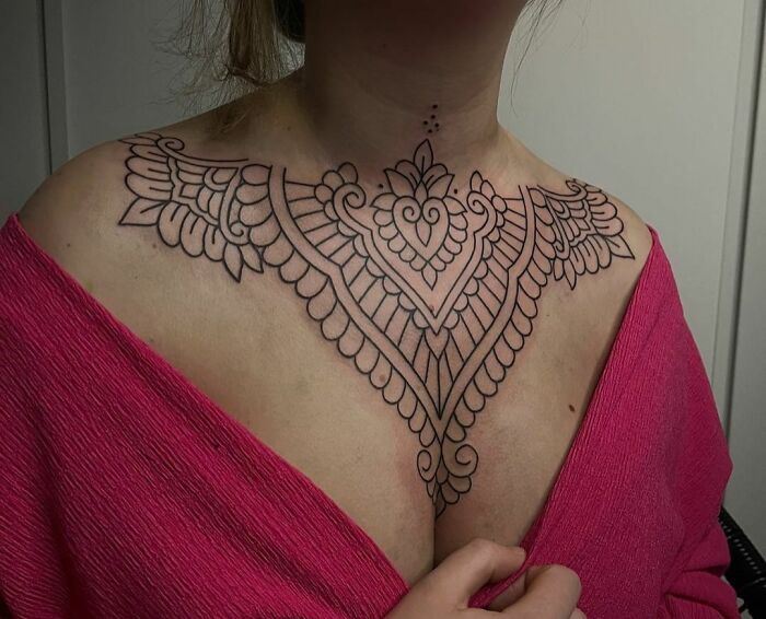 Intricate collarbone tattoo with symmetrical patterns on a woman's chest, wearing a pink top.