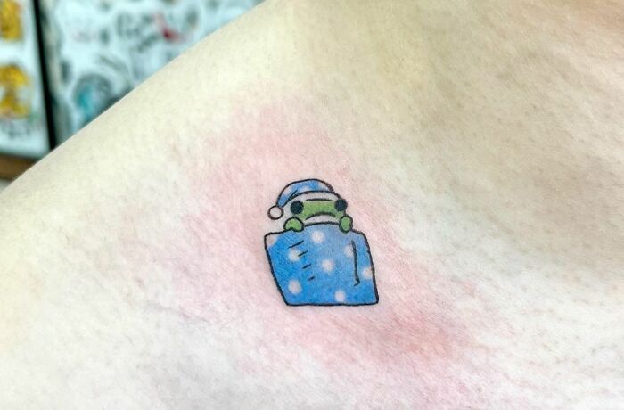 Collarbone tattoo of a cartoon frog in a blue blanket and nightcap.