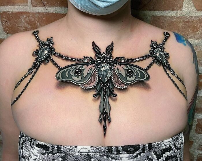 Intricate collarbone tattoo featuring a detailed moth design with chains connecting shoulder pieces.