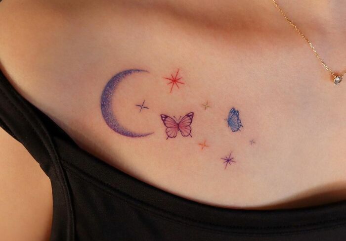 Colorful collarbone tattoo featuring a crescent moon, butterflies, and stars on a person wearing a black top.