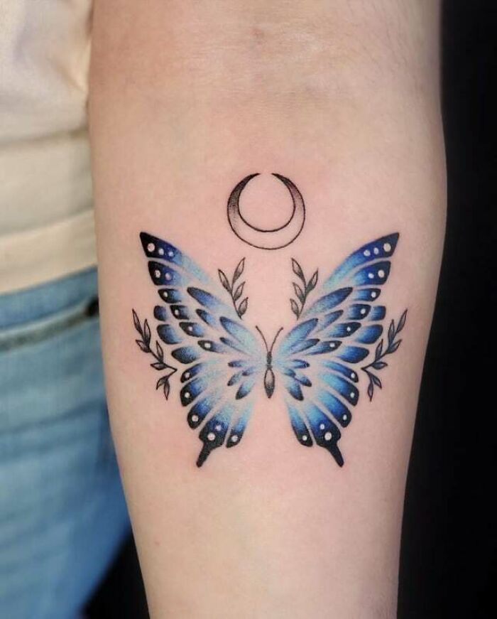 Colorful butterfly tattoo with crescent moon and leaf details on forearm.