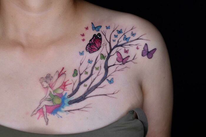 Colorful butterfly tattoo with tree branches and a fairy on a woman's shoulder.