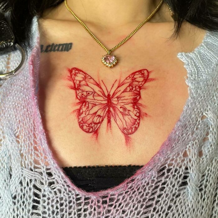 A red butterfly tattoo on a woman's chest, detailed and vibrant, under a pendant necklace.