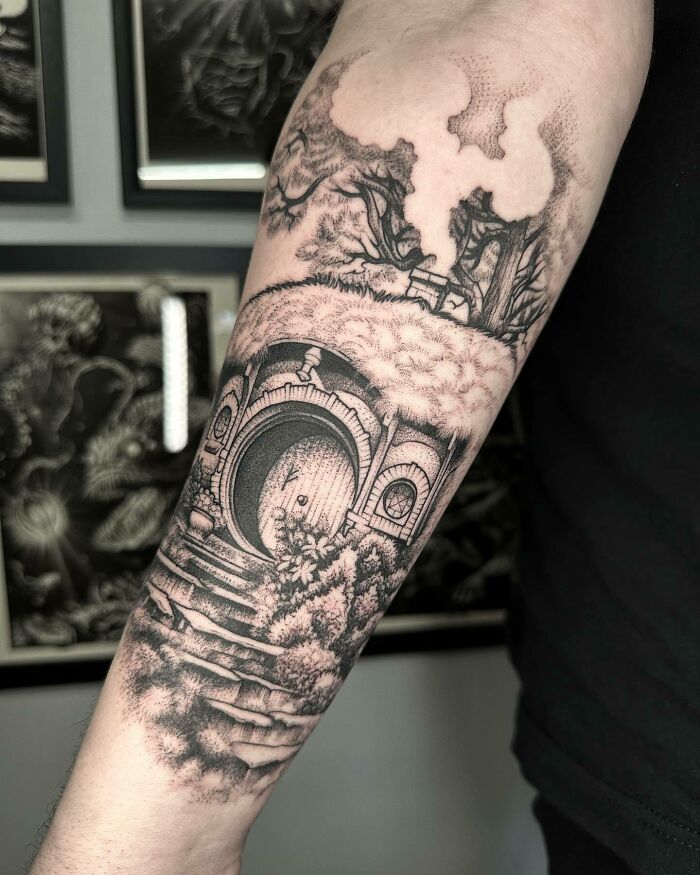 Detailed Lord of the Rings tattoo on forearm featuring a hobbit house with trees and a stone path.