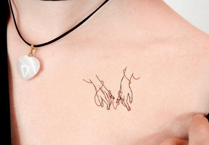 Minimalist collarbone tattoo of intertwined hands with a heart-shaped necklace above.