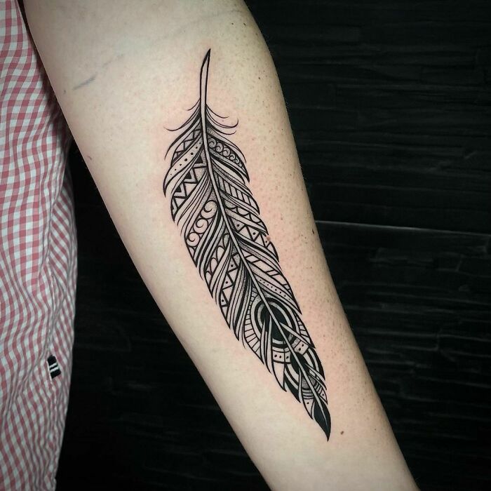 Tribal tattoo design of a detailed feather on a forearm against a dark background.