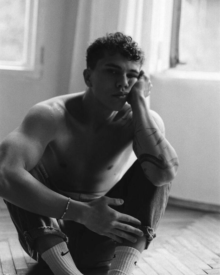 Shirtless man sitting on the floor, showcasing an arm tattoo.