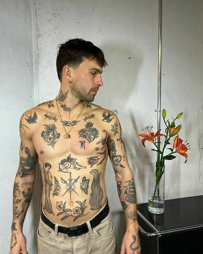 Man with diverse patchwork tattoos on his body, standing near a vase with bright orange lilies.