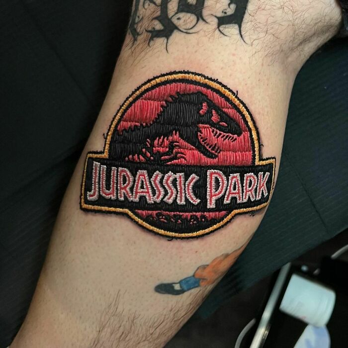 Pop culture tattoo of Jurassic Park logo on leg, featuring black and red embroidery-style design.
