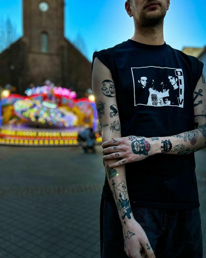 Man with patchwork tattoos on arms in front of a colorful carousel, wearing a black graphic tee.