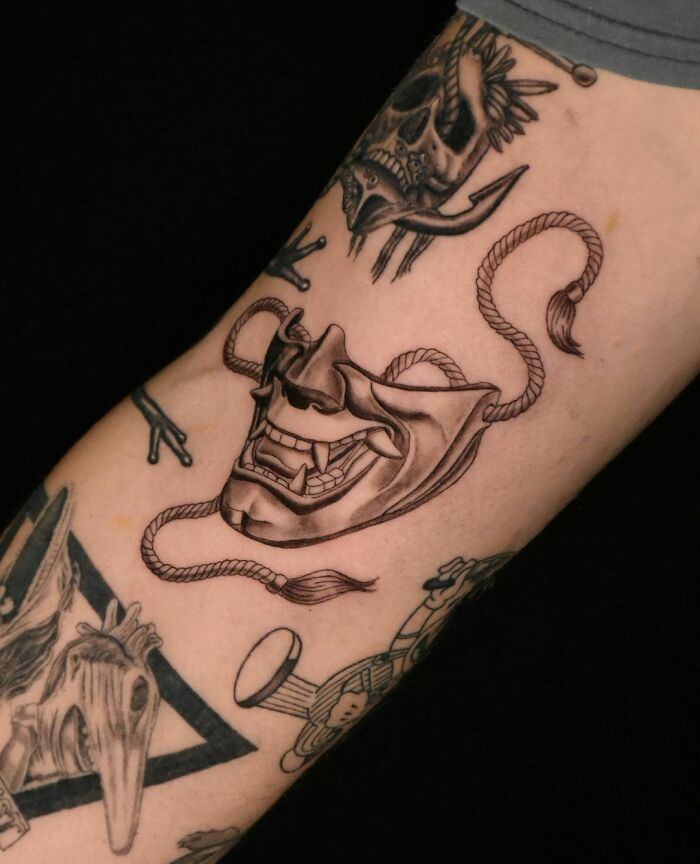 Patchwork tattoo with intricate black ink designs featuring skull and mask motifs on forearm.