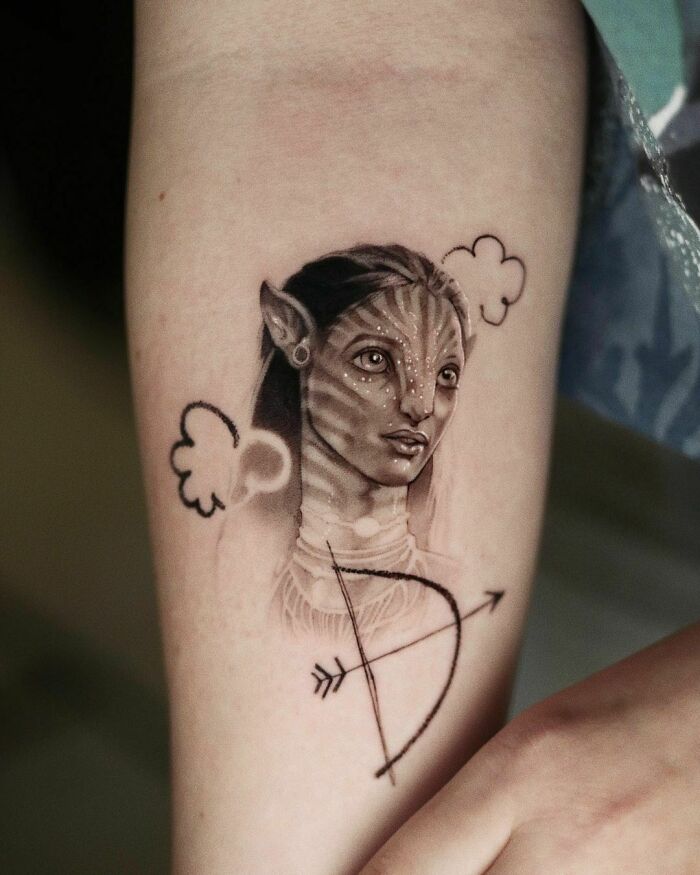 Tattoo of a Na'vi character from Avatar with cloud outlines and an arrow, representing pop culture inspiration.