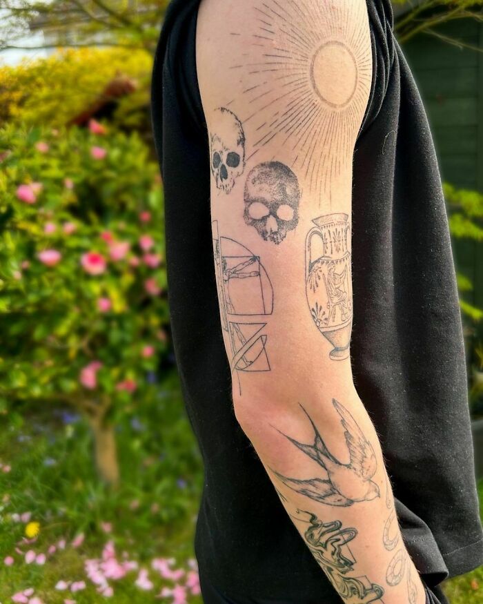 Arm featuring a patchwork tattoo design with skulls, a sun, and a bird, set against a garden background.