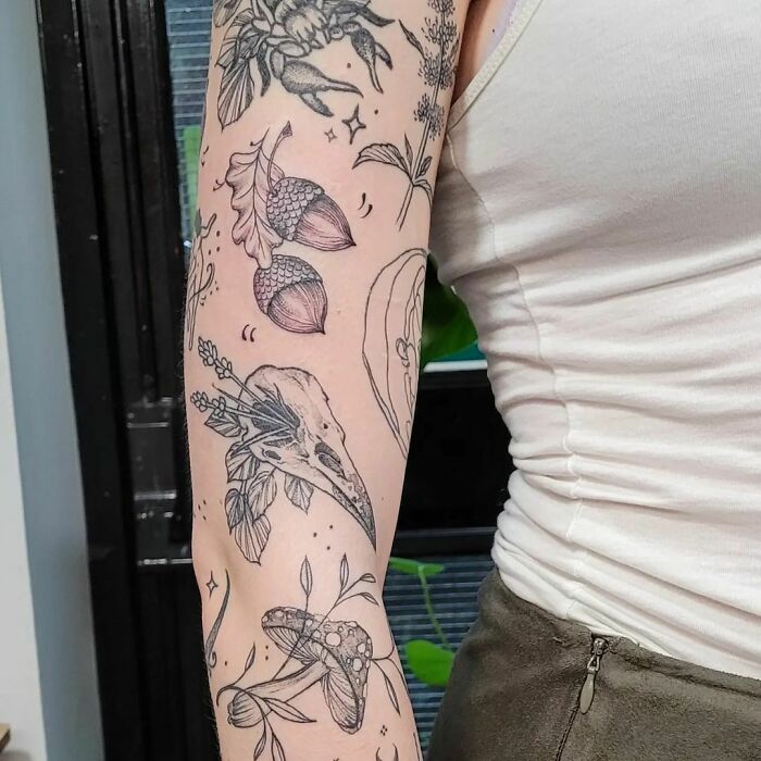Arm with a patchwork tattoo featuring nature-inspired designs like leaves, acorns, and a bird skull.