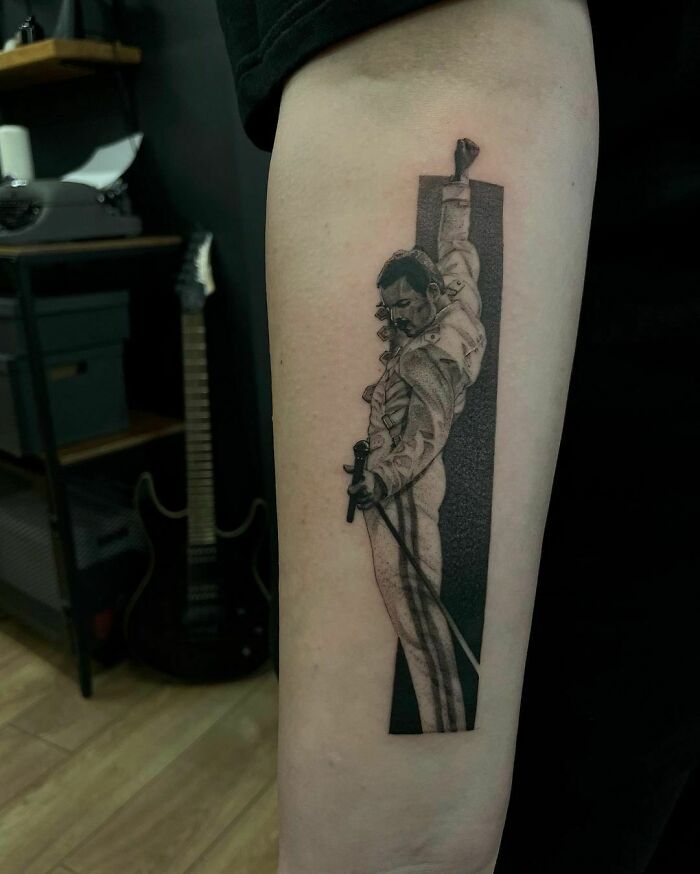 Pop culture tattoo of a performer in a dynamic pose on forearm, with a guitar in the background.