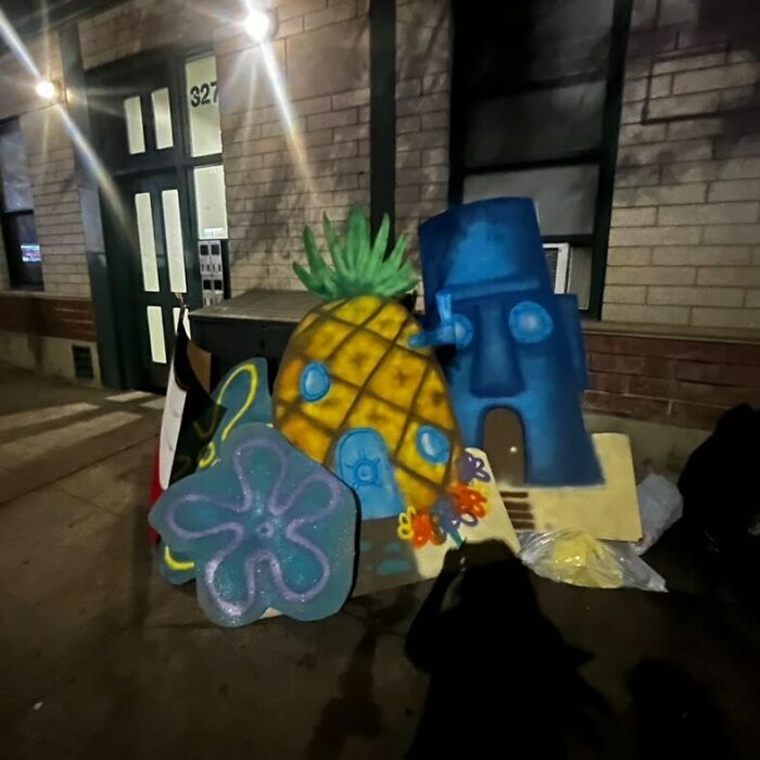 Colorful SpongeBob-themed graffiti art featuring a pineapple house and blue tiki face discarded on a NYC stoop at night.