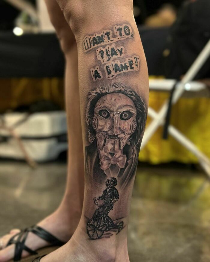 Pop culture tattoo featuring a ventriloquist dummy with text "Want to play a game?" on a person's leg.