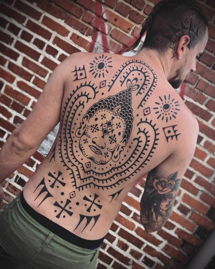 Man with intricate tribal tattoo on his back featuring geometric patterns and a face, standing against a brick wall.