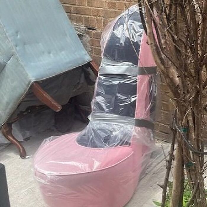Pink high-heeled shoe chair wrapped in plastic and taped, discarded next to brick wall and bare tree in NYC street.