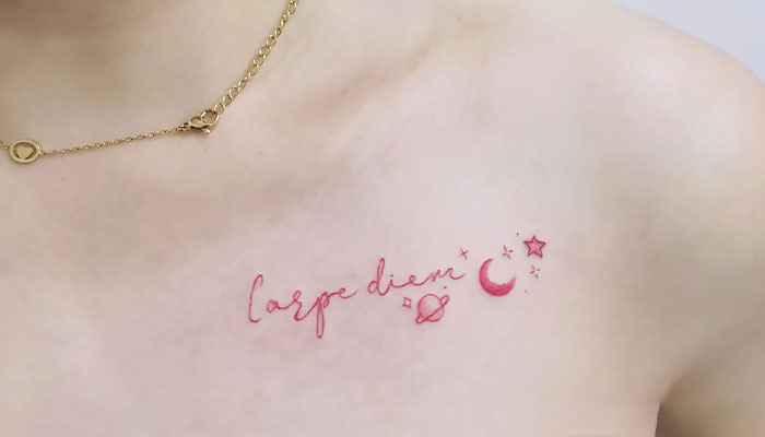 Collarbone tattoo with "carpe diem" in pink ink, adorned with stars, moon, and planet.
