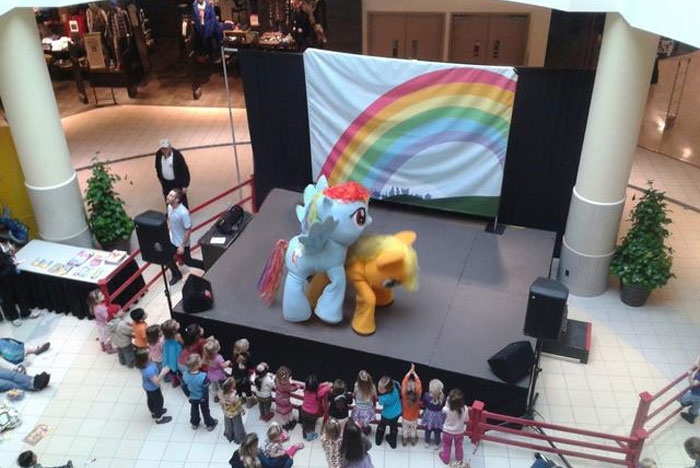 Cursed meme featuring My Little Pony characters in costume on stage at a mall with a rainbow backdrop and audience watching.