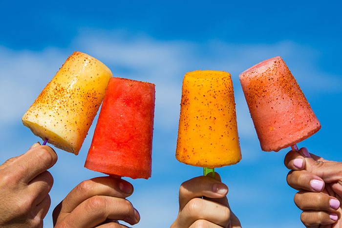 Four hands holding colorful popsicles against a blue sky, showcasing food history facts about sweet treats and what we eat.