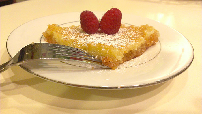 A slice of lemon bar with powdered sugar and two raspberries on top, showing the food history of desserts.