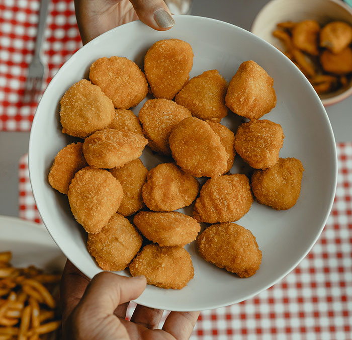 Hands holding a white plate full of golden-brown chicken nuggets, on a table with a red checkered cloth, showing food history facts.
