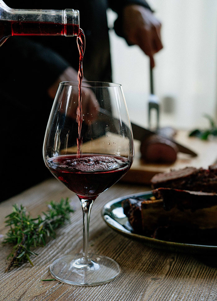 Red wine pouring into a glass, with a person carving meat in the background, representing food history and dining.