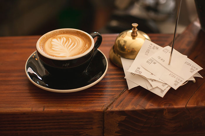 A cup of coffee with latte art next to a bell and stack of receipts, showcasing food history.