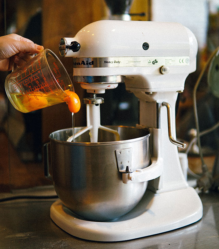 Food history facts: Eggs being poured from a measuring cup into a white stand mixer, detailing the story behind what we eat.