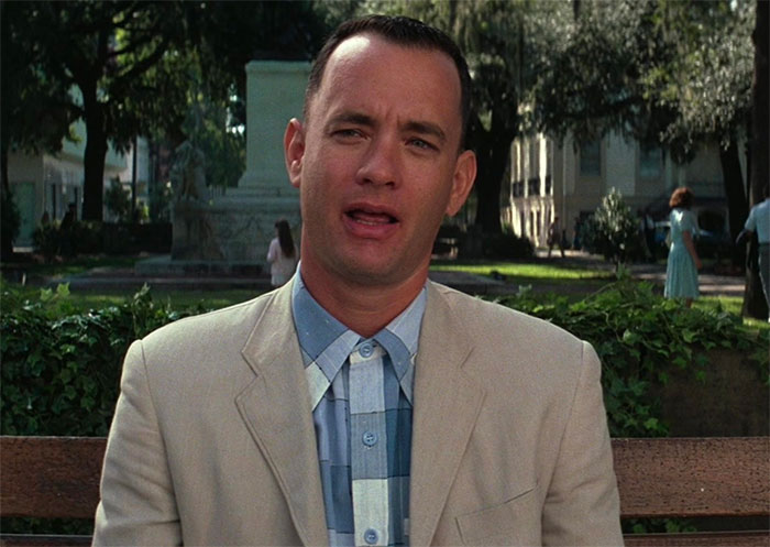 Forrest Gump sitting on a bench outdoors, wearing a beige jacket and blue checkered shirt from iconic Forrest Gump quotes scene