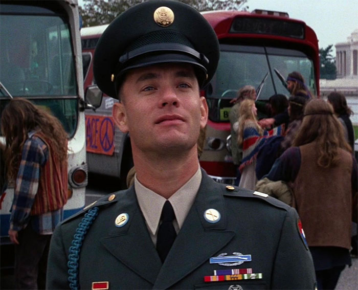Forrest Gump in military uniform standing outdoors with people and buses in the background, related to Forrest Gump quotes.