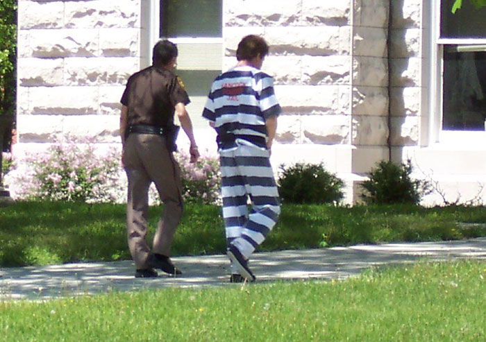 Prisoner in striped uniform being escorted by officer outside a building, representing disturbing secrets uncovered about someone close.
